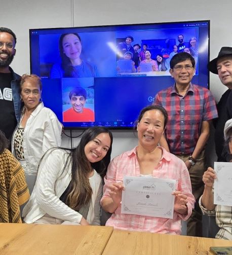 A group of people smiling for a graduation photo, with two participants holding certificates and others joining virtually on a screen in the background.