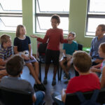 Child leading group storytelling in classroom setting, surrounded by peers and teacher.