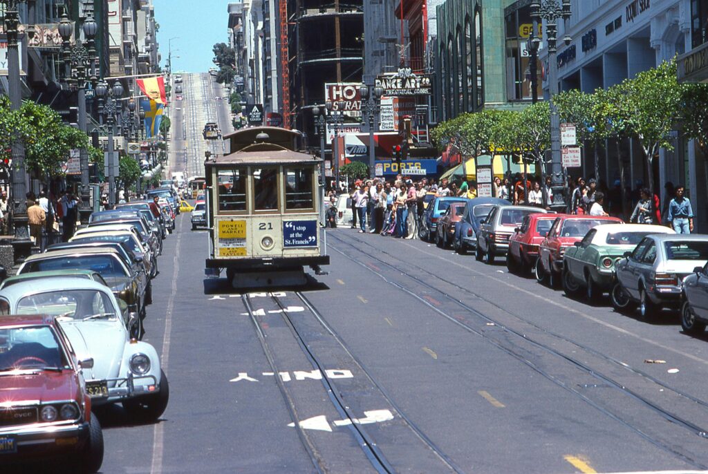 San Francisco cable car on Powell Street with classic cars and a crowd of people waiting on the sidewalk.