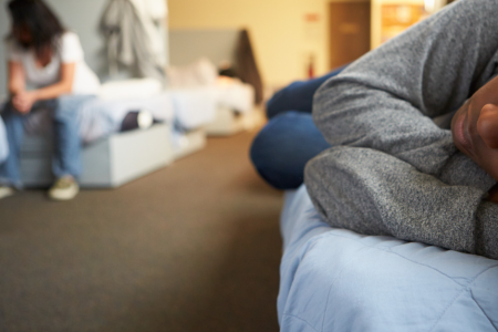 A person lying on a bed in a shelter with another individual sitting in the background, both in a shared room setting with minimal belongings and subdued lighting.